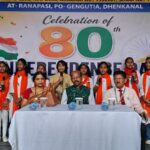 Schoolgirls in white outfits with orange stoles sing on stage beside a table of officials during an 80th independence day celebration banner in the background with the indian flag